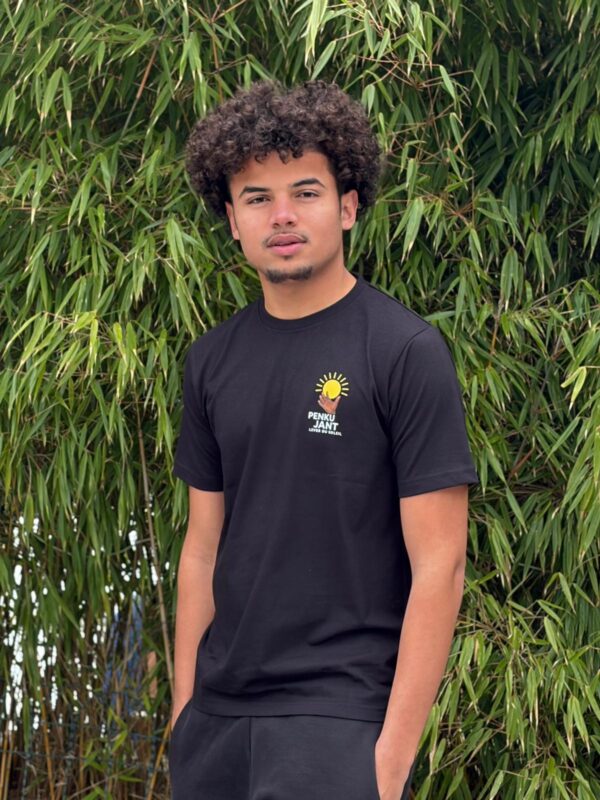 Junger Mann trägt schwarzes Tshirt und steht vor einem Baum.