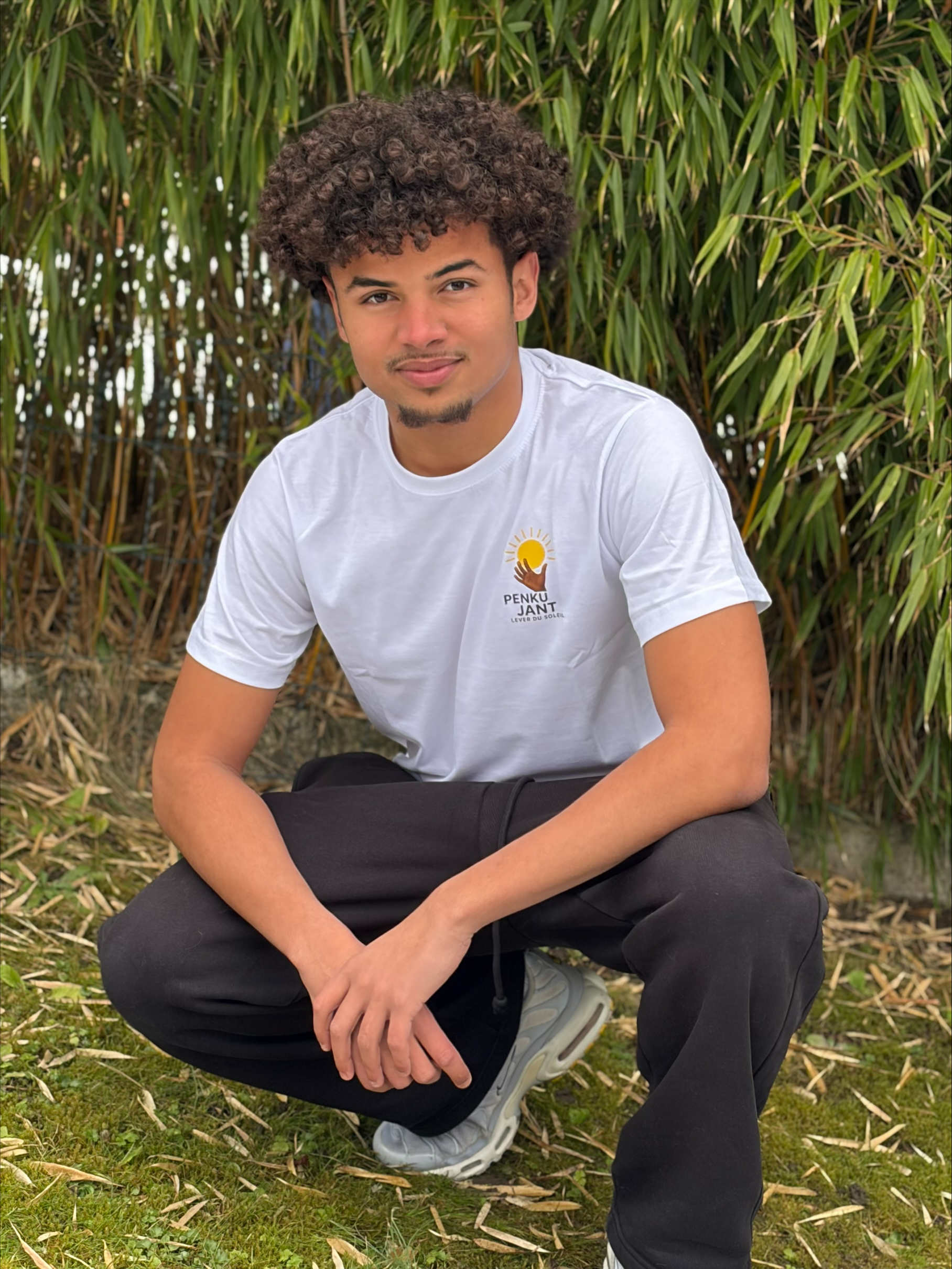 Junger Mann trägt weisses Tshirt und ist in der Hocke vor einem Baum.
